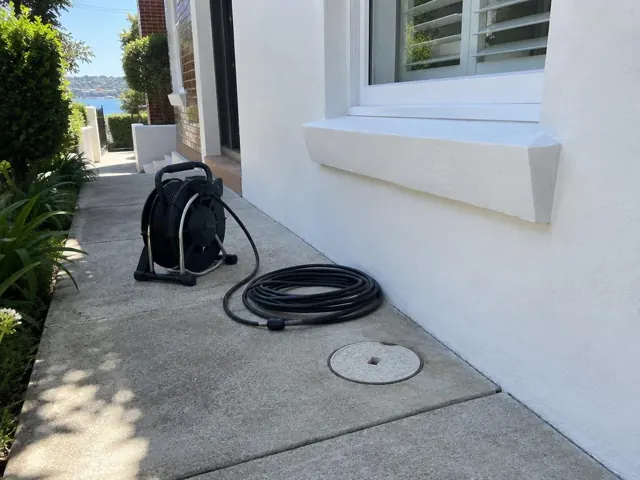 Concrete side path beside a white house with a capped inspection cover and coiled camera reel in Double Bay