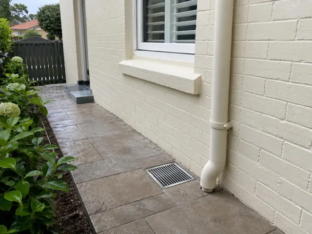 Outdoor gully grate beside cream brick wall and paved side path in Bellevue Hill