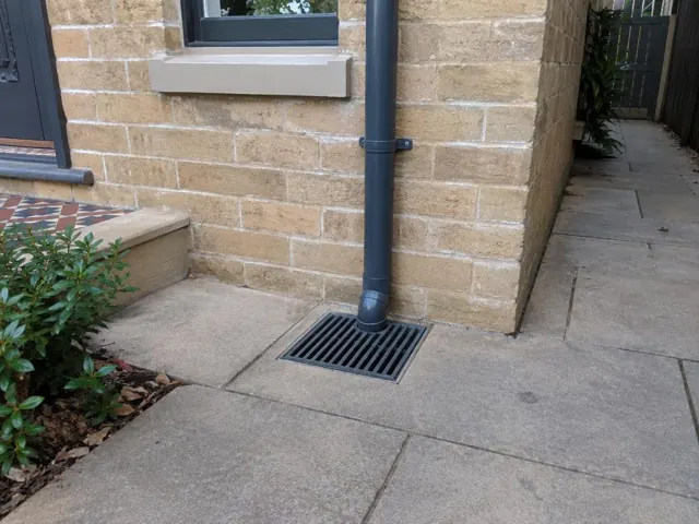 Stormwater grate beside a brick house wall with a downpipe and dry paving in Bondi